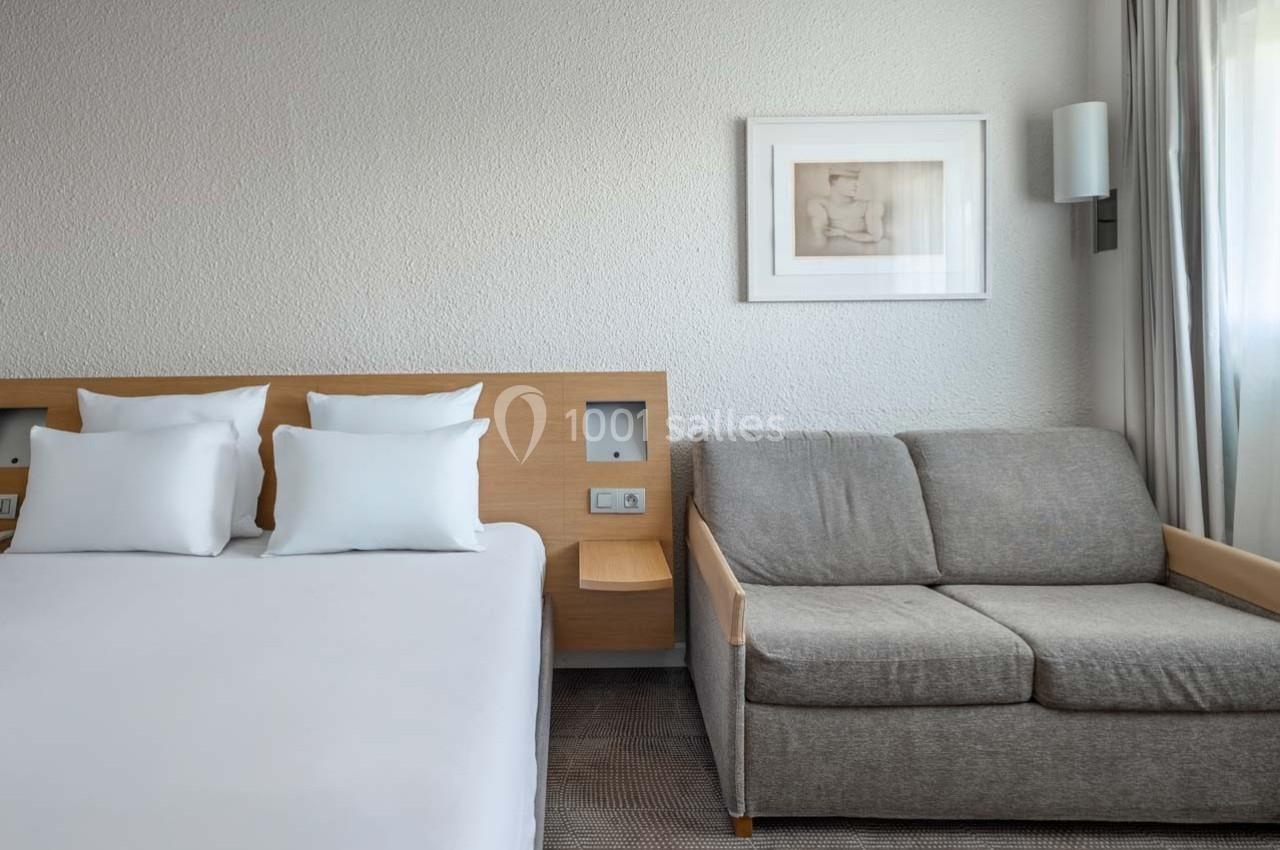 Chambre d'hôtel avec un lit double, un canapé gris, un mur blanc décoré d'un cadre et une lumière naturelle.