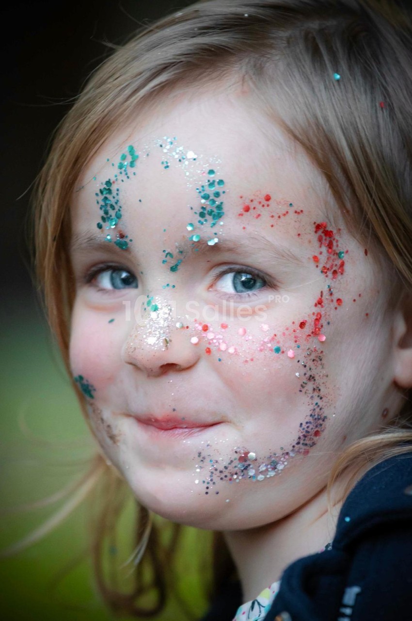 Fillette au visage maquillé de paillettes turquoise, rouges et argentées, regardant vers la droite sur un fond flou vert et sombre.