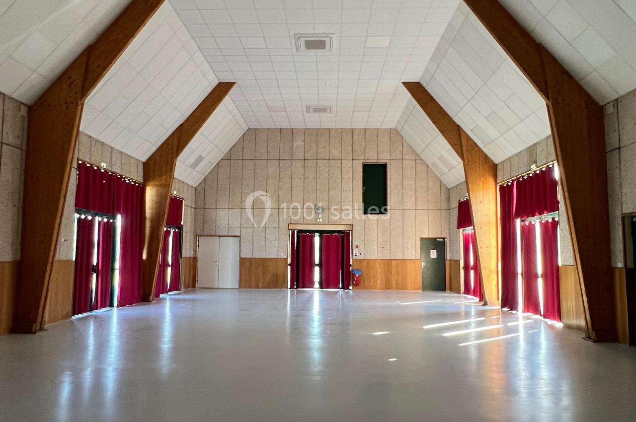 Salle vide avec un sol clair, des murs en bois et des rideaux rouges encadrant de grandes fenêtres.