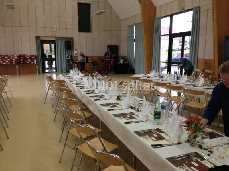 Salle de réception avec tables dressées pour un repas, nappes blanches, serviettes pliées et décorations florales.