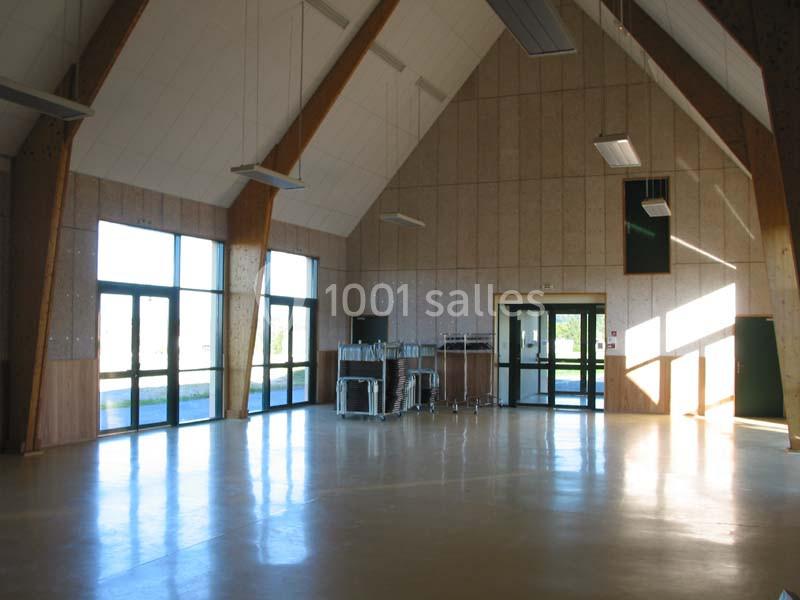 Salle spacieuse avec un sol clair, des murs en bois, de grandes fenêtres et des tables pliées rangées contre un mur.