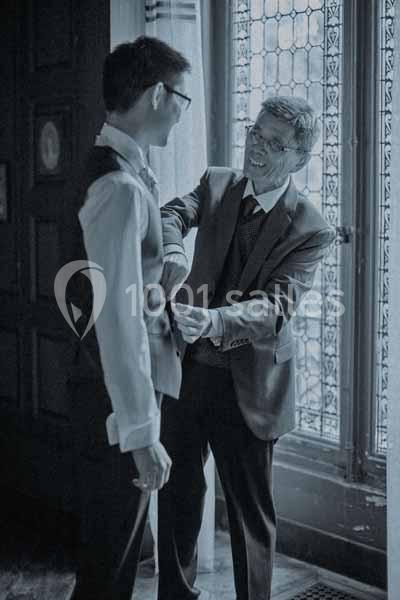 Un homme ajuste le gilet d'un jeune homme dans une pièce éclairée par une fenêtre à vitraux.