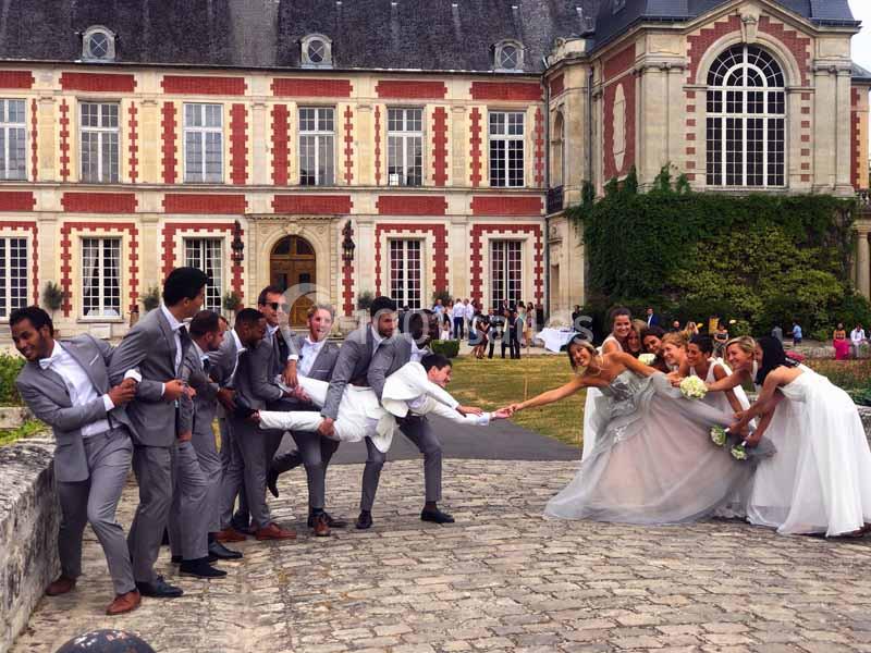 Des mariés et leurs invités en tenue de cérémonie jouent à tirer une corde devant un bâtiment historique.