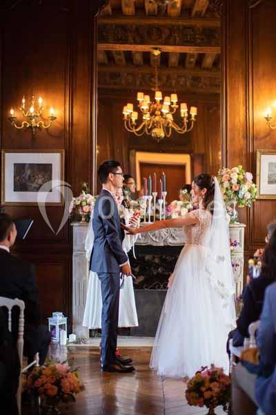 Un couple échange ses vœux de mariage dans une salle élégante avec boiseries, chandeliers et décor floral.