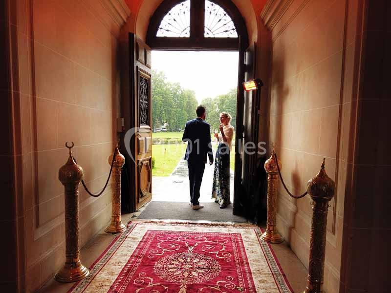 Un couple élégamment vêtu sort d'un bâtiment par une porte ouverte donnant sur un jardin verdoyant.