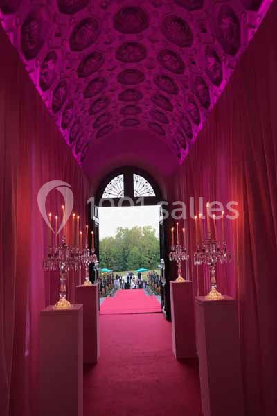 Allée décorée de chandeliers et de rideaux roses menant à une porte ouverte sur un espace extérieur.