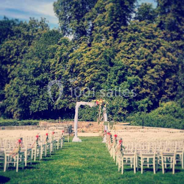 Chaises blanches alignées en extérieur devant une arche décorée, dans un jardin verdoyant sous un ciel dégagé.