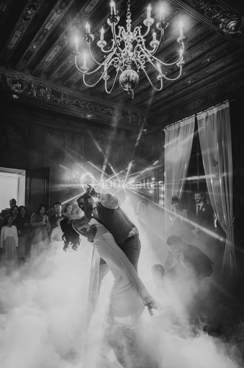 Un couple danse sous un lustre dans une salle sombre, entouré de fumée et d'invités observant la scène.