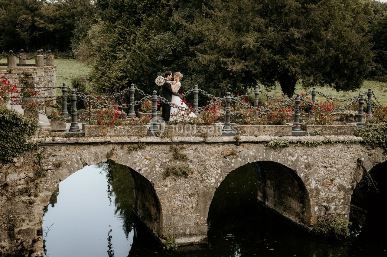 Un couple en tenue de mariage se tient sur un pont en pierre entouré de fleurs et de verdure.