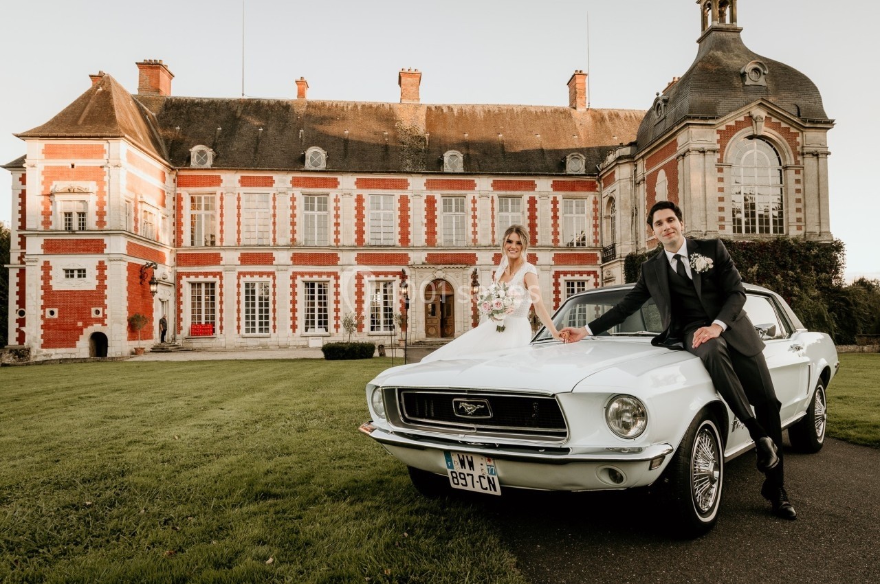 Un couple en tenue de mariage pose devant une voiture ancienne blanche, avec un château en arrière-plan.