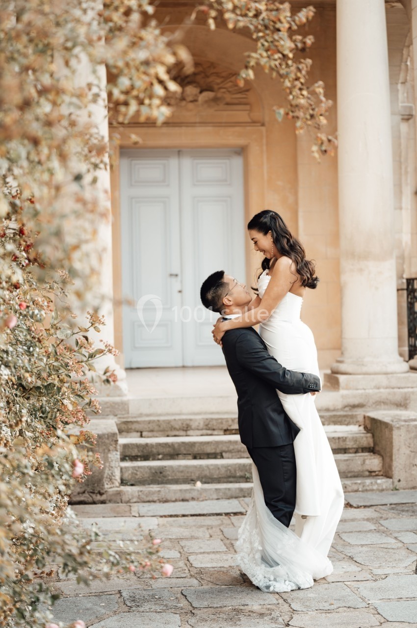 Un couple en tenue de mariage, l'homme portant la femme devant un bâtiment aux colonnes et une porte claire.