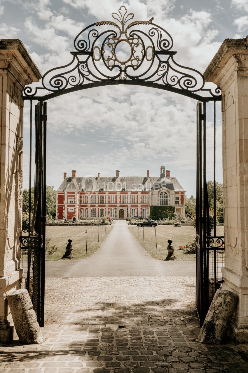 Portail en fer forgé ouvert donnant sur une allée menant à un château en pierre rouge entouré de verdure.