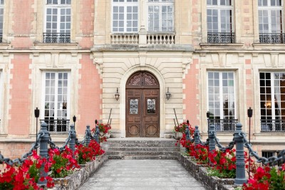 Miniature Location salle Lésigny (Seine-et-Marne) - Château de Lésigny #19 Façade d'un bâtiment ancien avec porte en bois sculpté, grandes fenêtres et allée bordée de fleurs rouges.