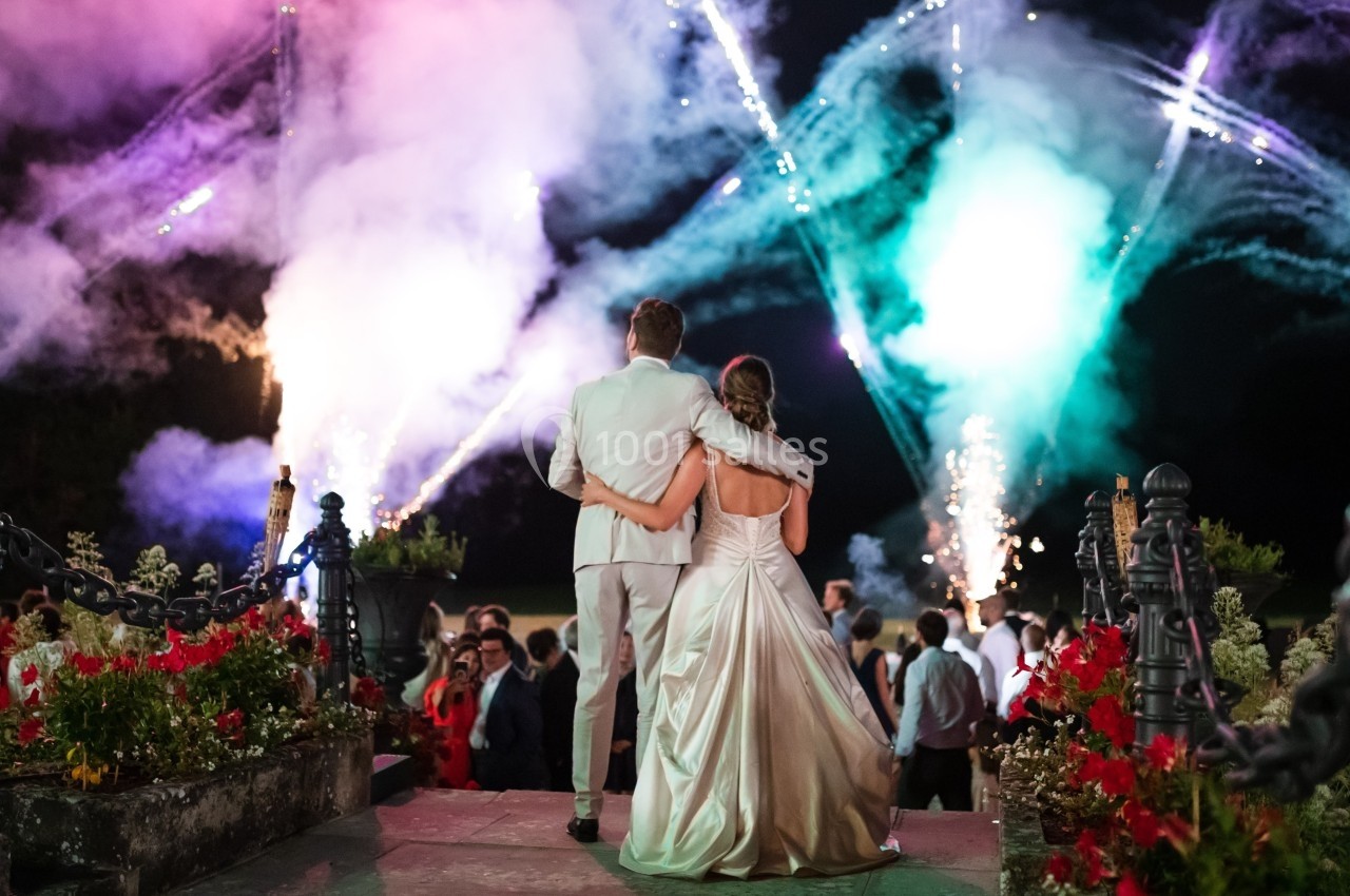 Un couple en tenue de mariage regarde un feu d'artifice coloré, entouré d'invités dans un jardin de nuit.