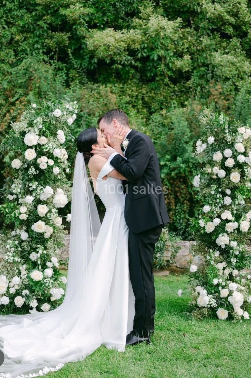 Un couple en tenue de mariage s'embrasse devant une arche florale blanche dans un jardin verdoyant.