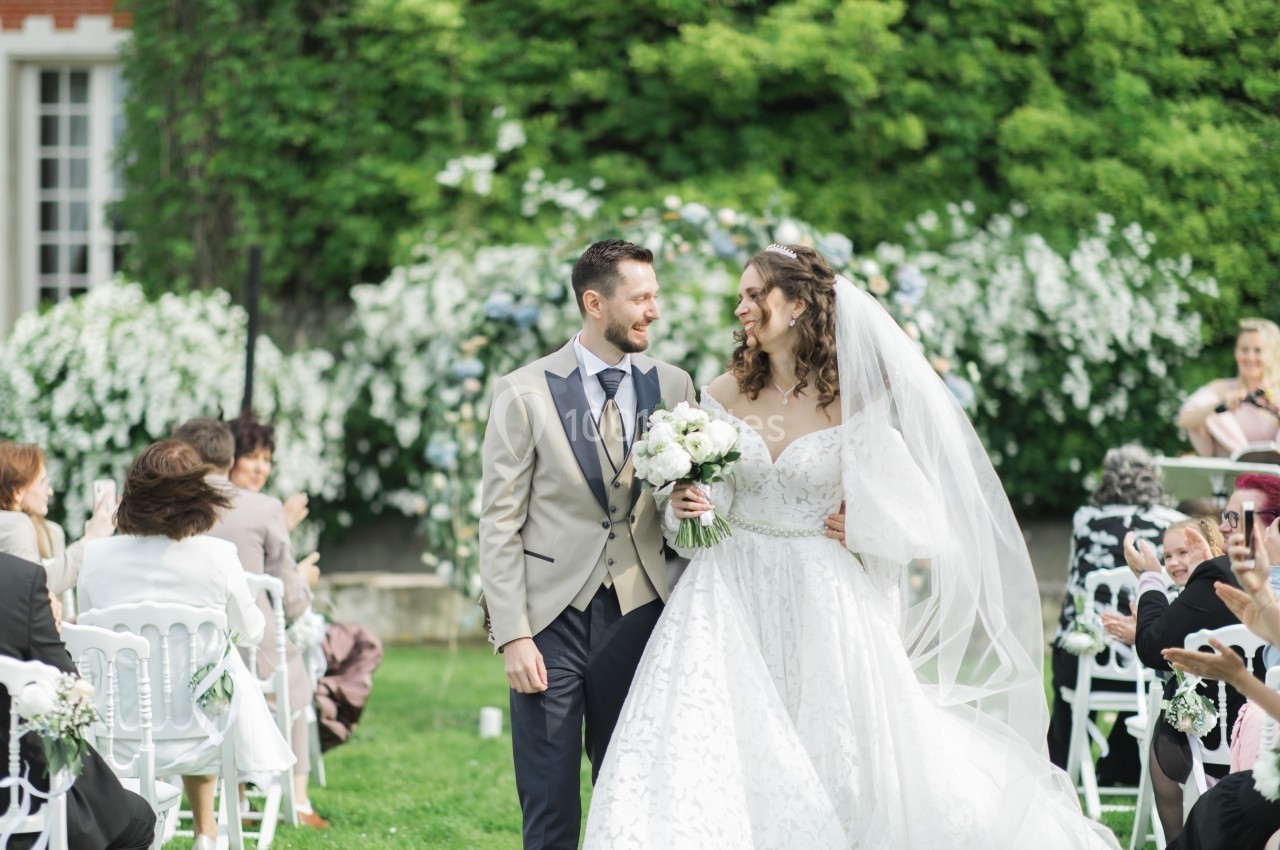 Un couple souriant marche dans une allée extérieure lors d'une cérémonie de mariage, entouré d'invités et de fleurs.