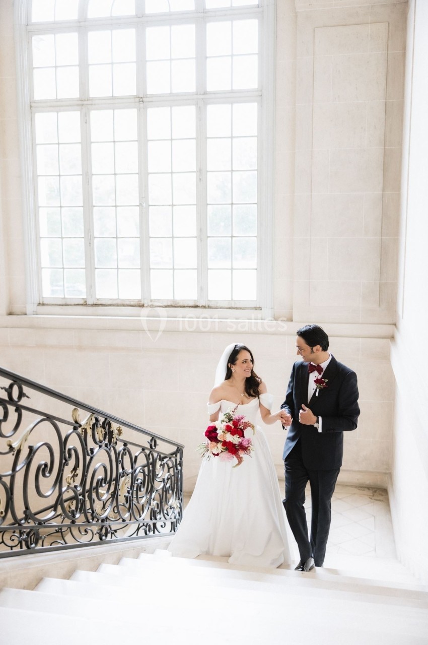 Un couple de mariés descend un escalier élégant dans un bâtiment lumineux avec une grande fenêtre en arrière-plan.