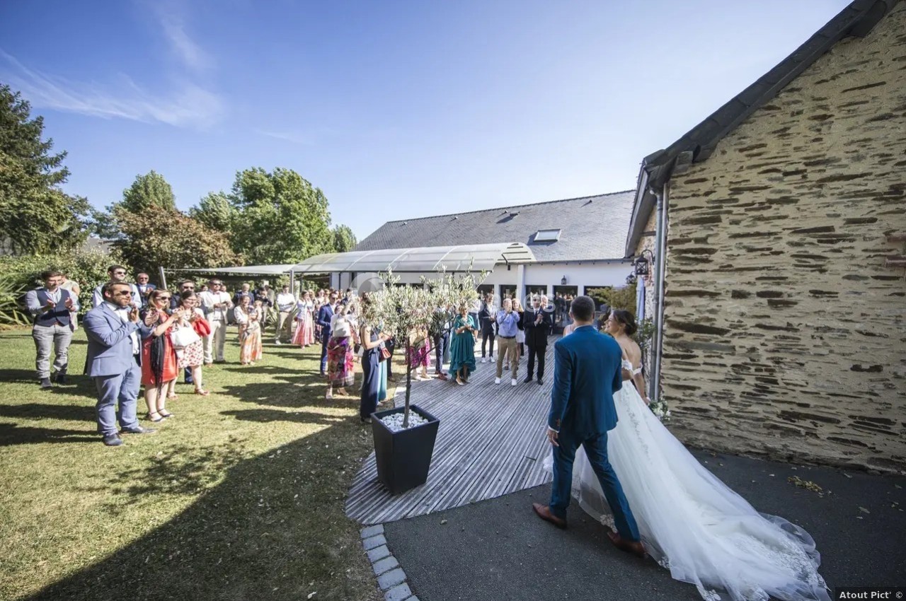 Un couple de mariés avance vers des invités rassemblés dans une cour extérieure près d'un bâtiment en pierre.