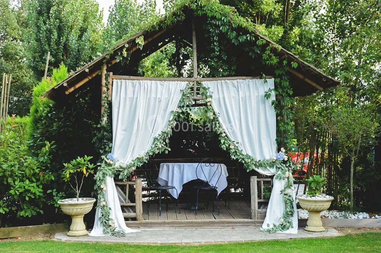 Abri en bois décoré de rideaux blancs et de feuillage, abritant une table dressée dans un jardin verdoyant.