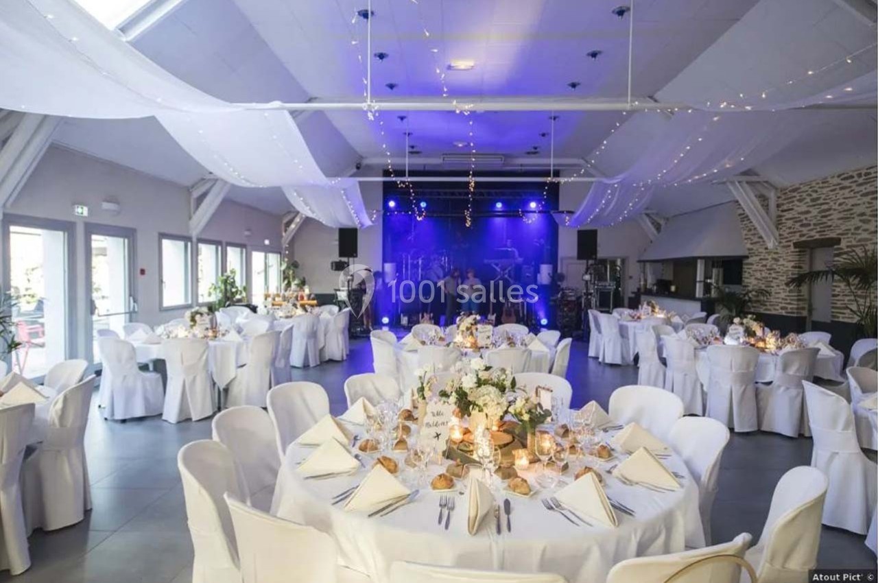 Salle de réception décorée avec des tables rondes dressées, éclairage tamisé et scène en fond avec projecteurs.