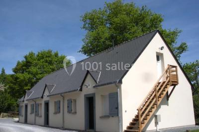 Cabane en bois avec fenêtres et porte vitrées, entourée de fleurs et d'arbustes, située près d'un chemin en gravier.