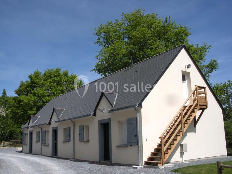 Maison à un étage avec toit en ardoise, volets bleus et escalier extérieur en bois, entourée d'arbres.