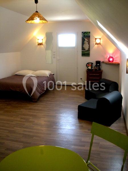 Chambre mansardée avec lit double, fauteuils noirs, table verte, parquet et éclairage chaleureux.