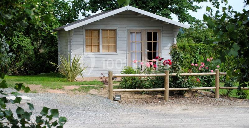 Cabane en bois avec fenêtres et porte vitrées, entourée de fleurs et d'arbustes, située près d'un chemin en gravier.