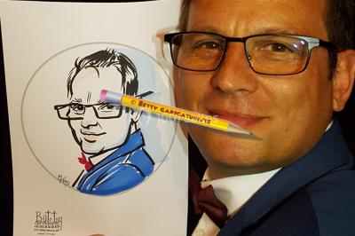 Un homme souriant tient un dessin caricatural de lui-même, réalisé avec des détails humoristiques.