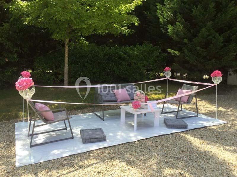 Salon de jardin décoré avec des rubans roses, des coussins gris et des fleurs, installé sur un tapis blanc en extérieur.
