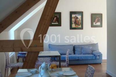 Salle à manger avec table dressée, chaises transparentes, poutre apparente, canapé bleu et tableaux au mur.