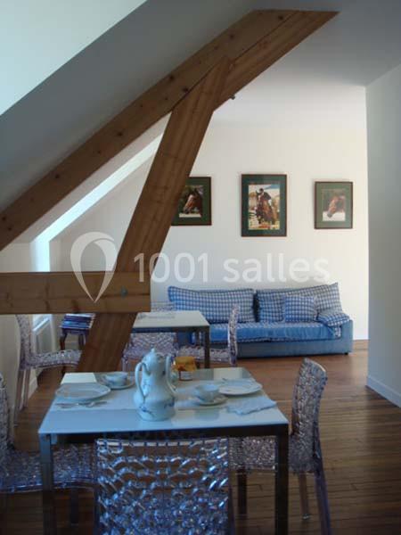 Salle à manger avec table dressée, chaises transparentes, poutre apparente, canapé bleu et tableaux au mur.