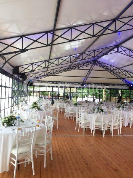 Salle de réception lumineuse avec tables rondes dressées, chaises blanches et structure métallique apparente.