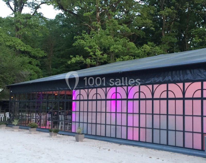 Grande tente avec parois vitrées et éclairage rose à l'intérieur, située dans un environnement boisé.