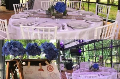 Salle à manger avec table dressée, chaises transparentes, poutre apparente, canapé bleu et tableaux au mur.