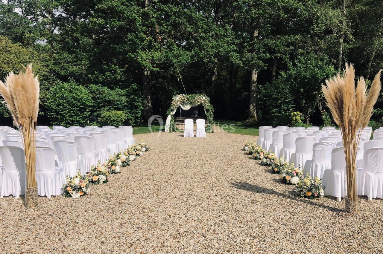 Allée centrale bordée de chaises blanches et de fleurs menant à une arche décorée, dans un cadre extérieur verdoyant.