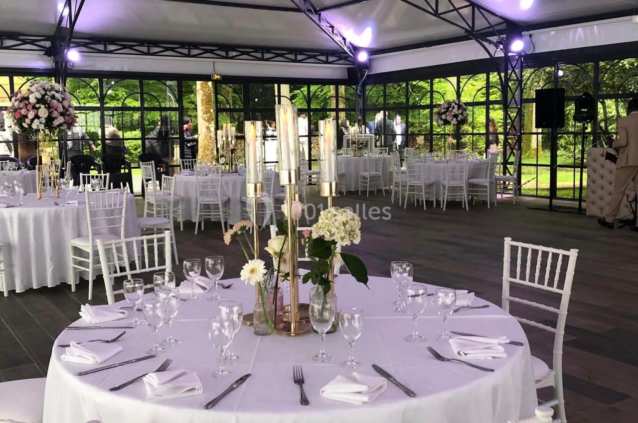 Salle de réception sous une structure en toile avec tables rondes dressées, décor floral et lumière naturelle.