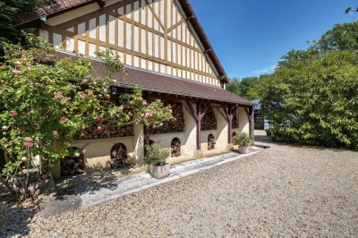 Façade d'un bâtiment à colombages avec portes ouvertes, donnant sur une cour en gravier et des arbres en arrière-plan.