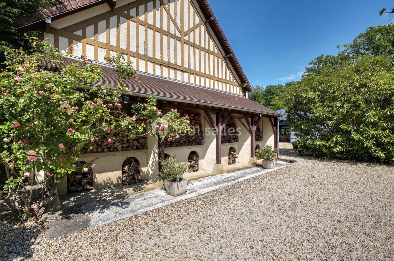 Façade d'une maison à colombages avec un toit en pente, rangée de bûches et arbustes fleuris devant.