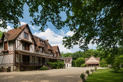 Façade d'un bâtiment à colombages avec portes ouvertes, donnant sur une cour en gravier et des arbres en arrière-plan.
