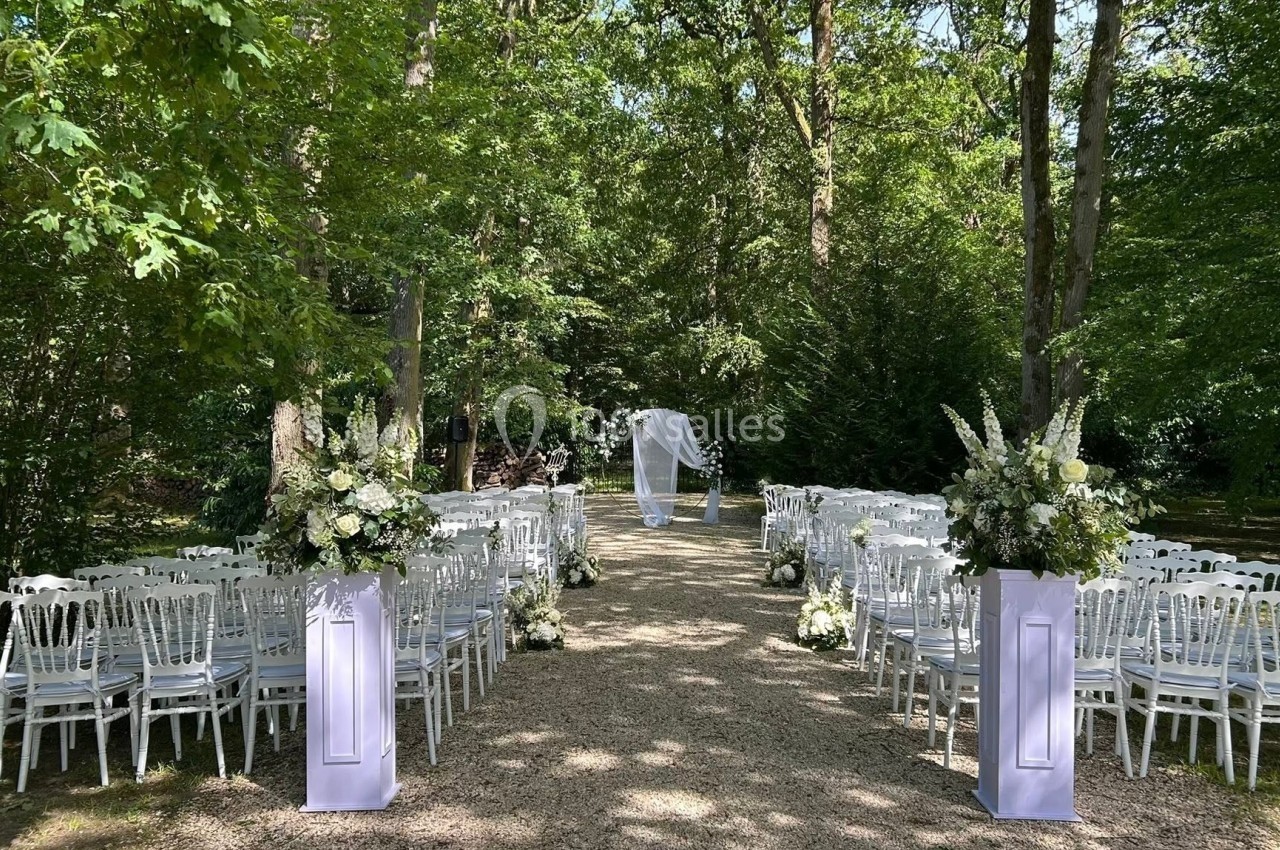 Allée centrale bordée de chaises blanches et décorations florales, menant à une arche de mariage dans un jardin boisé.