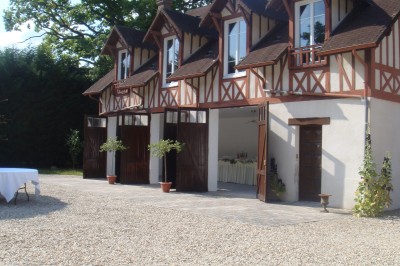 Façade d'un bâtiment à colombages avec portes ouvertes, donnant sur une cour en gravier et des arbres en arrière-plan.