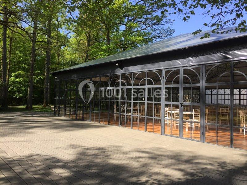 Structure en verre et métal avec des tables et chaises à l'intérieur, entourée d'arbres et d'une terrasse en bois.