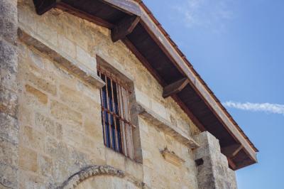 Façade en pierre d'un bâtiment ancien avec une fenêtre à barreaux et un toit en bois sous un ciel bleu.