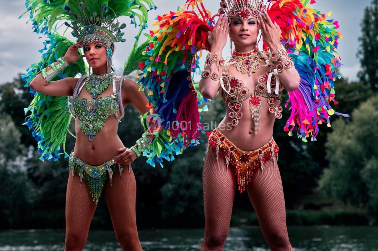 Deux danseuses en costumes colorés avec plumes, posant devant un plan d'eau et des arbres en arrière-plan.