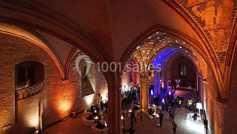 Vue intérieure d'une grande salle voûtée illuminée par des lumières colorées, avec des invités et des espaces aménagés.