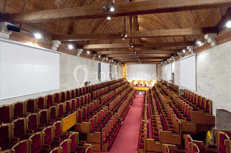 Salle de conférence avec rangées de sièges en bois et coussins rouges, plafond voûté en bois et murs en pierre.