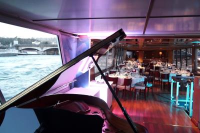 Salle de restaurant sur un bateau avec tables rondes dressées, nappes blanches et vue sur un paysage fluvial.