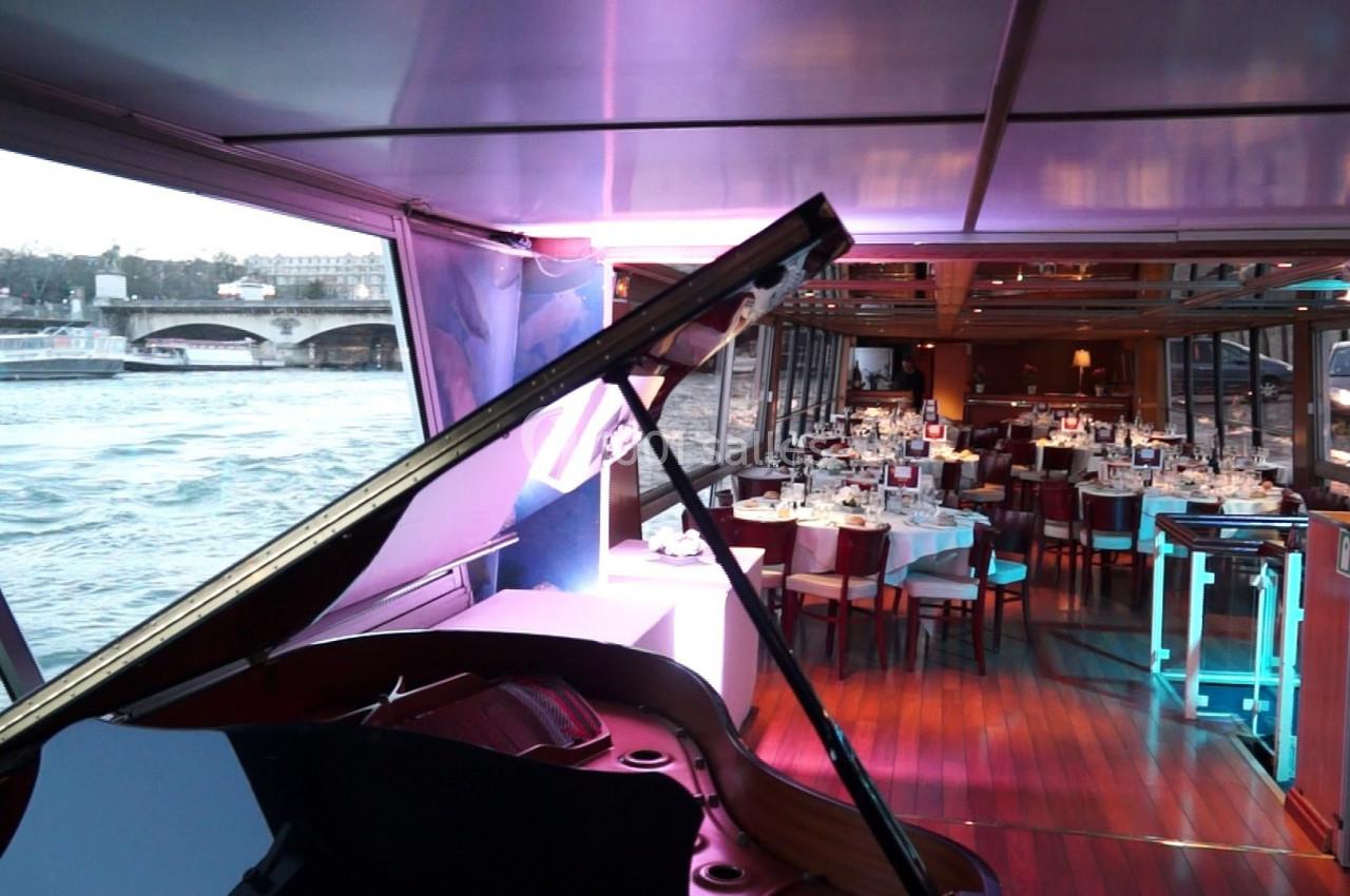 Vue intérieure d'un bateau avec un piano à queue au premier plan et une salle de restaurant élégamment dressée en arrière…