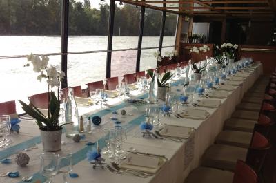 Salle de restaurant sur un bateau avec tables rondes dressées, nappes blanches et vue sur un paysage fluvial.